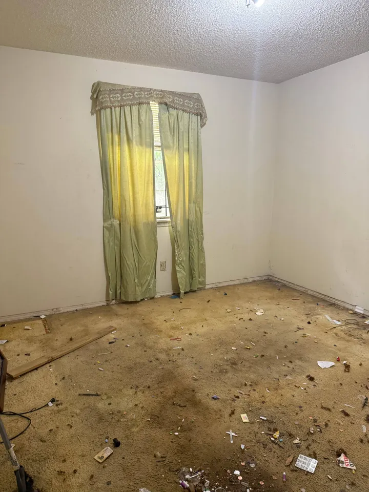 An empty bedroom with faded green curtains and heavily soiled carpet covered in small debris and rubbish.