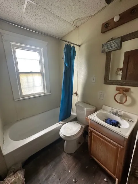 A dated bathroom with a stained tub, a blue shower curtain, a wood-framed mirror and worn flooring.