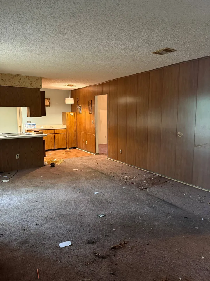 An empty living room with dark wood panelling, a dated kitchen beyond it, and worn carpet marked with stains and scattered debris.